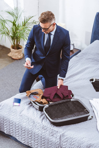 vue grand angle de l'homme en costume et lunettes à l'aide d'un smartphone et valise d'emballage pour le voyage d'affaires
 - Photo, image