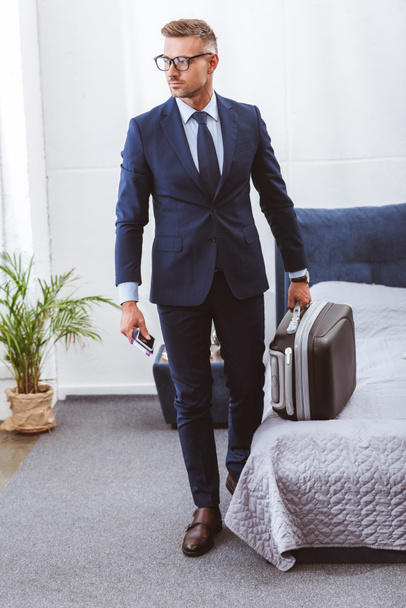 handsome businessman holding suitcase, smartphone and passport in bedroom - Photo, Image