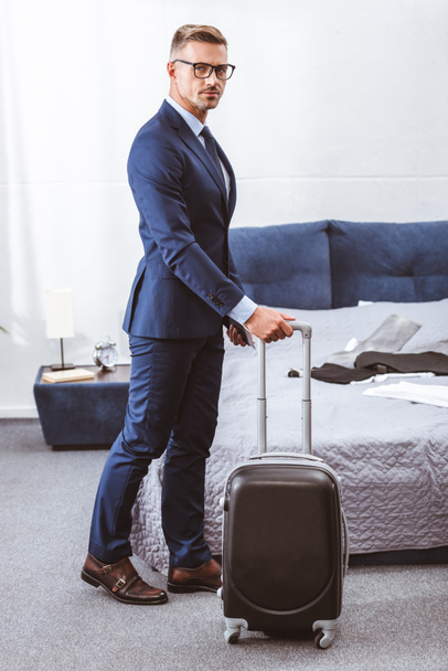 businessman in suit and eyeglasses holding suitcase and looking at camera in bedroom - Photo, Image