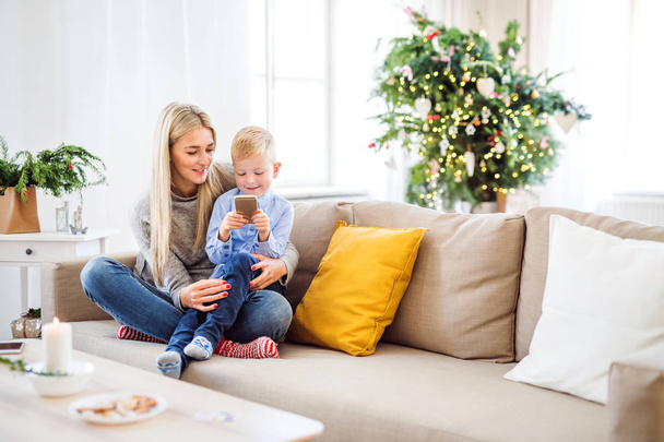 A mother and small boy with smartphone sitting on a sofa at home at Christmas time. - Foto, immagini