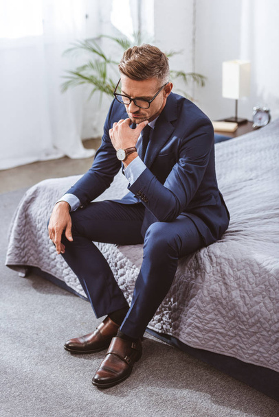 thoughtful man in suit and eyeglasses sitting on bed at home - Photo, Image