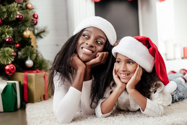 cheerful african american family in santa claus hats lying on floor in decorated room for christmas at home - Photo, Image