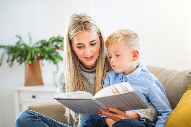 A mother and small boy sitting on a sofa at home at Christmas time, reading a book. - Foto, Imagen