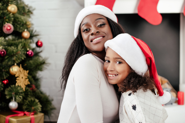 Portret van gelukkig Afro-Amerikaanse moeder en dochter in santa claus hoeden in ingerichte kamer thuis - Foto, afbeelding