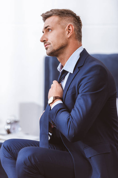 side view of handsome man in suit sitting and wearing necktie at home - Фото, зображення