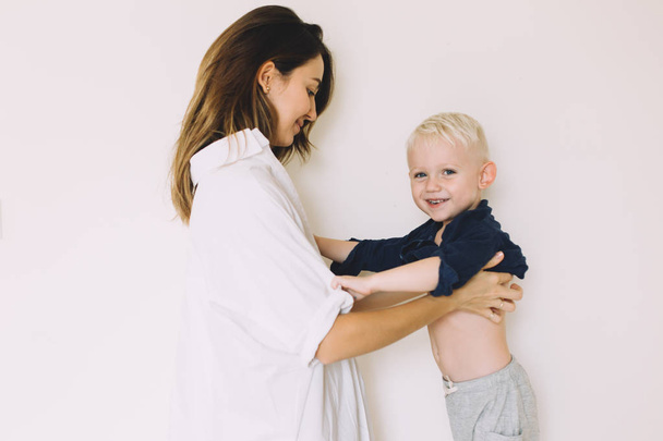 Young beautiful mother buttoning up the shirt of her little son in the bedroom - Fotoğraf, Görsel