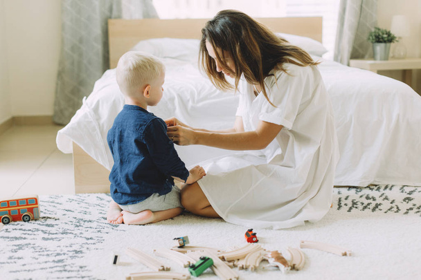 Young beautiful mother buttoning up the shirt of her little son in the bedroom - 写真・画像