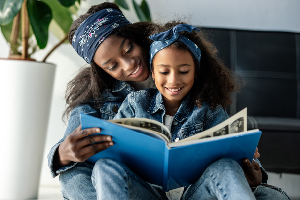 portrait of smiling african american mother and daughter looking at family photo album at home - Photo, Image
