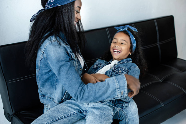 happy african american woman having fun with daughter in similar clothing on sofa at home - Foto, imagen