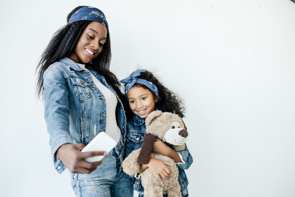 Lächelnde Afroamerikanerin und Tochter in ähnlicher Kleidung machen Selfie auf Smartphone - Foto, Bild