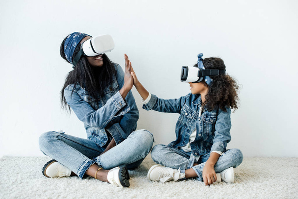 african american mother and daughter in similar clothing and vr headsets giving high five to each other at home - Foto, Bild