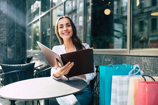 Young woman sitting in street cafe and reading menu after shopping - Photo, Image