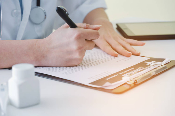 young female doctor or pharmacist with stethoscope writing prescription on clipboard  and bottle of pills on desk in hospital, health care, medical, medicine, pharmacy and insurance concept - Photo, image