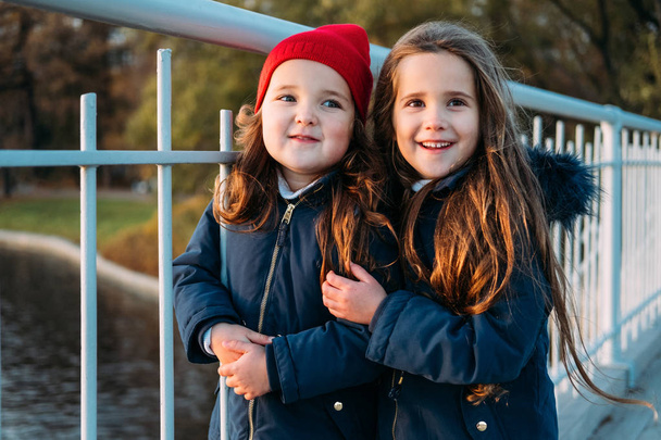 Két boldog gyermekek átölelve őszi parkban. Közelről napos életmód divat portréja két gyönyörű kaukázusi lányok szabadban, aranyos divatos ruhát visel. Little sisters nevetve, pihentető, élvezni az életet - Fotó, kép