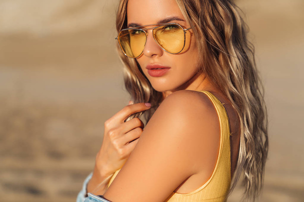 Close-up portrait of a sexy girl in a yellow swimsuit and sunglasses on the beach. Amazing woman face with make-up and beautiful hairstyle. Model on vacation, travels and sunbathes. - 写真・画像