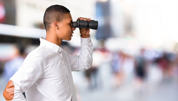 Young african american man with white shirt and looking for something in the distance with binoculars in the middle of the city - Photo, Image