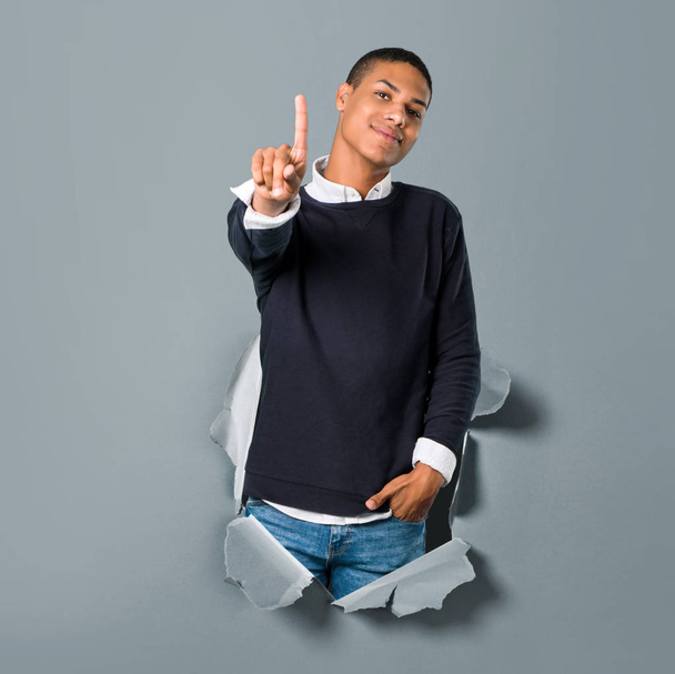 Νεαρός αφρικανική αμερικανική άνθρωπος που δείχνει και την άρση ένα δάχτυλο στο σημάδι από τα καλύτερα μέσα από μια τρύπα του χαρτιού - Φωτογραφία, εικόνα