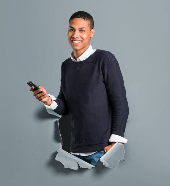 Young african american man looking at the camera and smiling while using the mobile through a paper hole - Фото, изображение