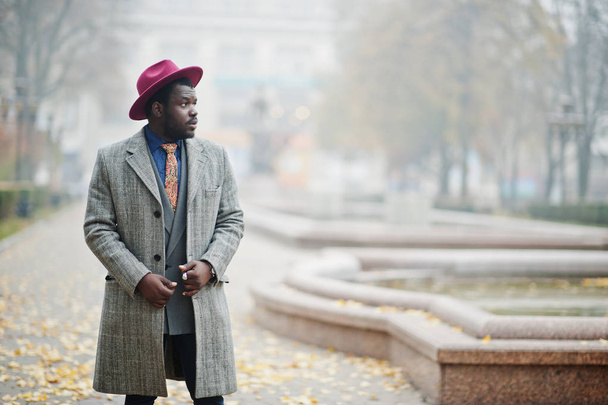 Stylish African American man model in gray coat, jacket tie and red hat posed at foggy weather street. - Foto, Bild