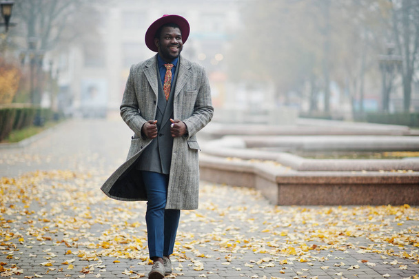 Stylish African American man model in gray coat, jacket tie and red hat posed at foggy weather street. - Foto, imagen