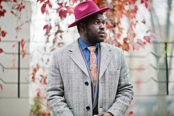 Stylish African American man model in gray coat, jacket tie and red hat posed against red leaves at autumn. - Foto, imagen
