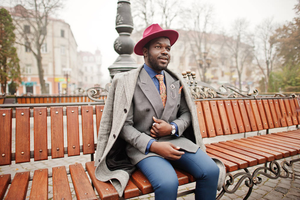 Stylish African American man model in gray coat, jacket tie and red hat posed at bench. - Foto, Imagen