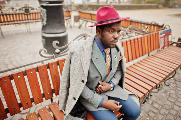 Stylish African American man model in gray coat, jacket tie and red hat posed at bench. - Foto, Imagem