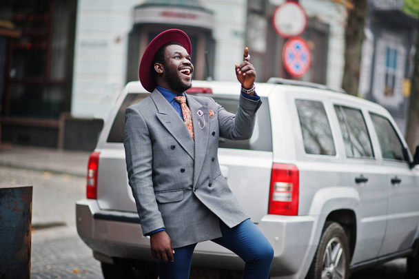 Elegante modelo de hombre afroamericano en corbata chaqueta gris y sombrero rojo posado contra suv coche de plata
. - Foto, Imagen