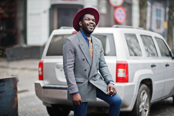 Stylowe African American man model w szarą kurtkę krawat i red hat postawione przed srebrny samochód suv. - Zdjęcie, obraz