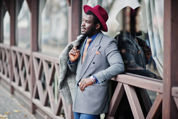 Stylish African American man model in gray coat, jacket tie and red hat posed against wooden cafe. - Photo, Image