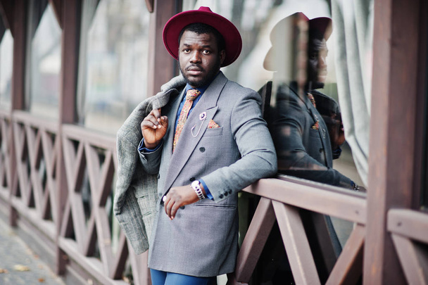 Stylish African American man model in gray coat, jacket tie and red hat posed against wooden cafe. - Фото, зображення
