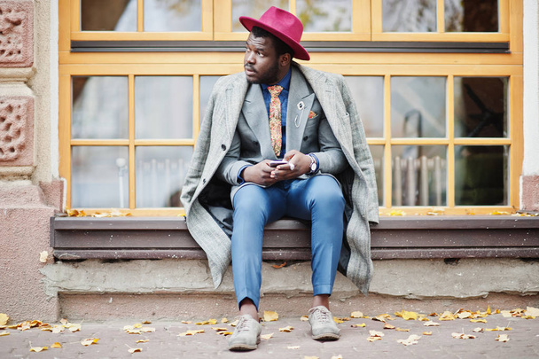 Stylish African American man model in gray coat, jacket tie and red hat sitting against large windows and holding phone at hands. - Foto, Bild