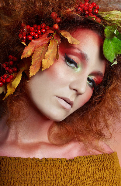 Τέχνης πορτρέτο του φθινοπώρου γυναίκες στα μαλλιά, ζωηρό πτώση χρώματα και μακιγιάζ, κόκκινα σγουρά μαλλιά και ογκώδη μαλλιά. Φύλλα και μούρα από ένα δέντρο σορβιά στο κεφάλι και στους ώμους του κοριτσιού - Φωτογραφία, εικόνα
