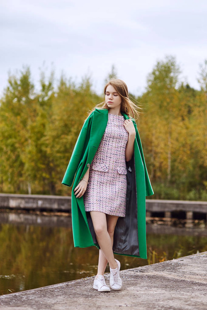 Girl in a green coat walks along the lake embankment on a cloudy autumn day. Autumn fashion and clothing, yellow fallen leaves floating in the water. Romantic mood - Foto, imagen