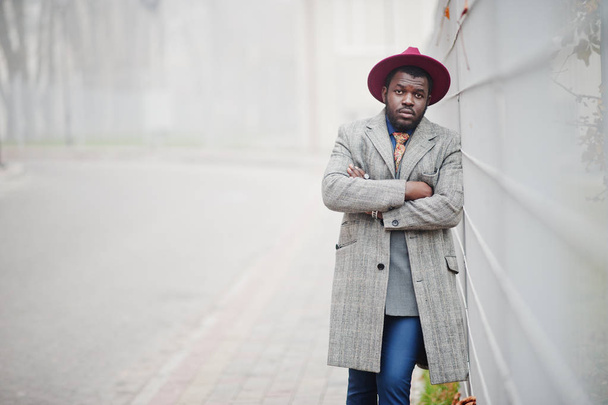 Stylish African American man model in gray coat, jacket tie and red hat against fence in fog day. - Photo, Image