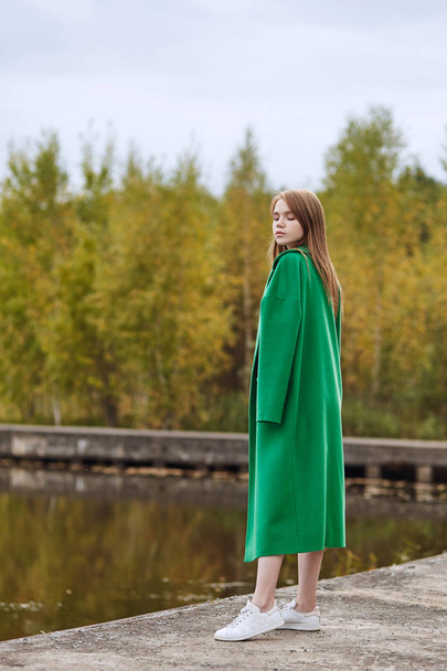 Girl in a green coat walks along the lake embankment on a cloudy autumn day. Autumn fashion and clothing, yellow fallen leaves floating in the water. Romantic mood - Photo, image