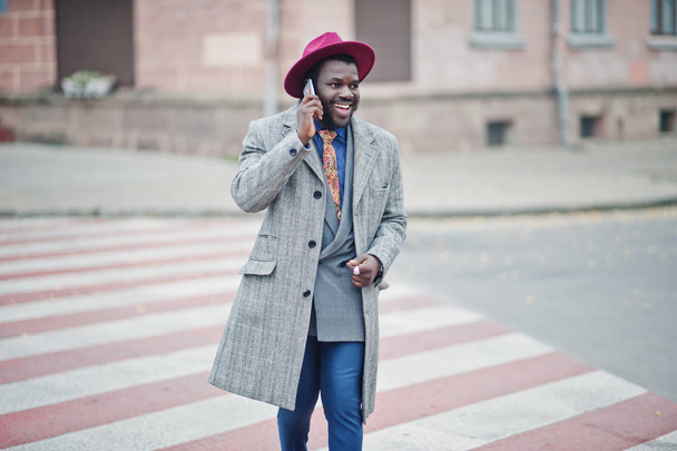 Stylish African American man model in gray coat, jacket tie and red hat walking on crosswalk and speaking on mobile phone. - Фото, изображение