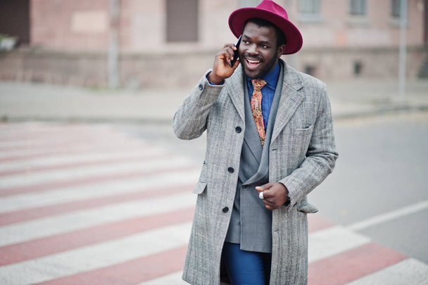 Stylish African American man model in gray coat, jacket tie and red hat walking on crosswalk and speaking on mobile phone. - Fotoğraf, Görsel