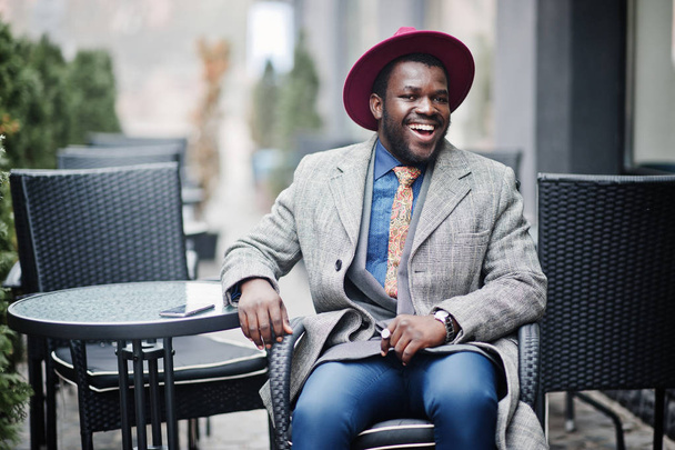 Κομψό αφρικανική αμερικανική άνδρα μοντέλο σε γκρι παλτό, μπουφάν γραβάτα και κόκκινο καπέλο sitiing στην εξωτερική καφετέρια. - Φωτογραφία, εικόνα