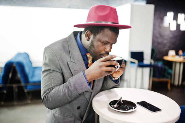 Stylish African American man model in gray jacket tie and red hat drink coffee at cafe. - Foto, imagen