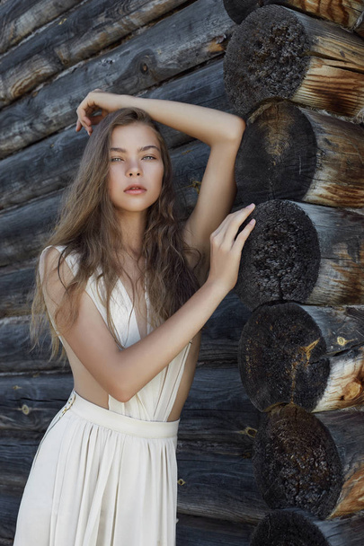 Sexy young woman in a light white dress posing in the village near the old house. Girl with a perfect figure on the background of a country house of logs - 写真・画像