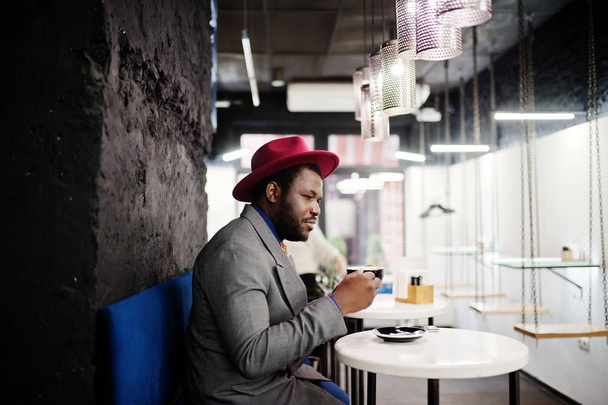 Stylish African American man model in gray jacket tie and red hat drink coffee at cafe. - Фото, зображення