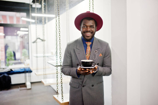 stylisches afrikanisch-amerikanisches Männermodel in grauer Sakko-Krawatte und rotem Hut trinkt Kaffee im Café. - Foto, Bild