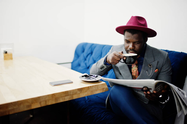 Stylish African American man model in gray jacket tie and red hat drink coffee at cafe and read newspapers. - Fotografie, Obrázek