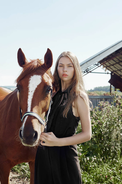 Girl rider stands next to the horse in the field. Fashion portrait of a woman and the mares are horses in the village in the grass. Blonde woman holding a horse by the bridle, beautiful body - Foto, Imagen