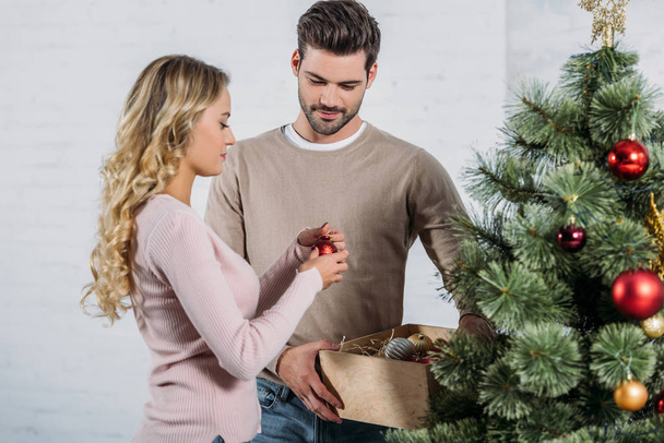 couple decorating christmas tree with baubles together at home, boyfriend holding wooden box with toys - Photo, Image