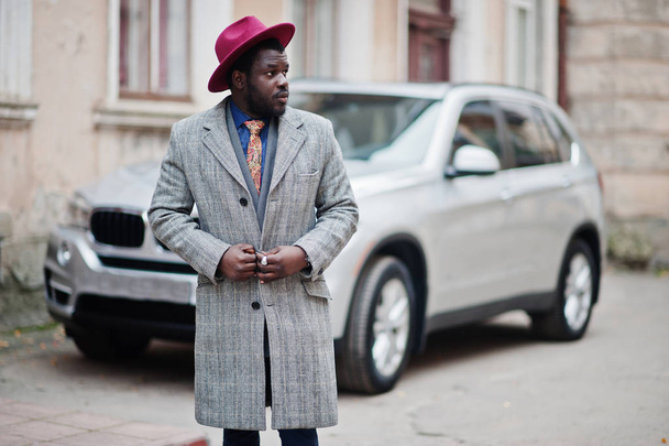 Stylish African American man model in gray coat, jacket tie and red hat against silver business suv car. - Foto, Imagem