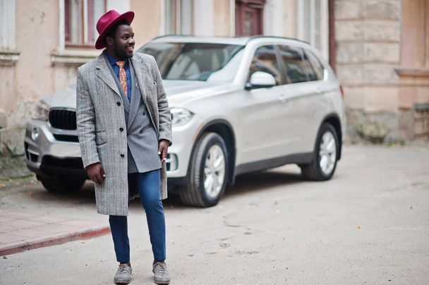 Stylish African American man model in gray coat, jacket tie and red hat against silver business suv car. - Foto, imagen