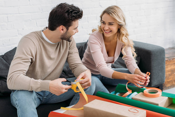 smiling couple preparing christmas gift box together, holding ribbons and looking at each other at home - Photo, Image