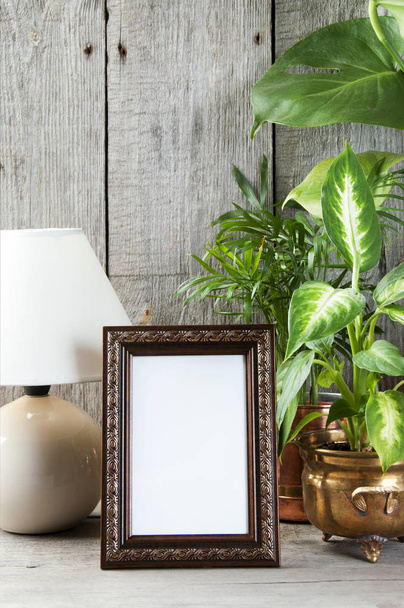 Üres barna függőleges képkeret, zöld növények és egy asztali lámpa a régi fa szürke textúrázott háttérre. Otthoni dekoráció és másol adható szöveg.   - Fotó, kép